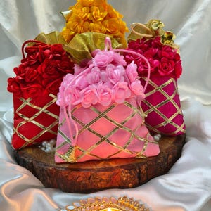 May include: An arrangement of colorful, decorative pouches with floral accents and gold trim. The pouches are red, pink, and yellow, and are arranged on a wooden surface. A gold candle holder with a lit candle is in the foreground.
