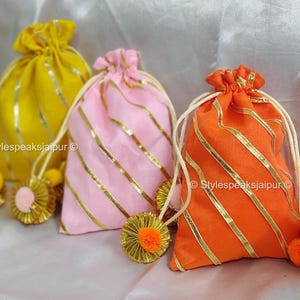Può includere: Tre sacchetti con coulisse gialli, rosa e arancioni, ciascuno decorato con strisce dorate e un nappa abbinata. I sacchetti sono legati con cordoncini bianchi e poggiano su una superficie bianca e morbida. Il testo "Stylespeaksjaipur" è visibile.