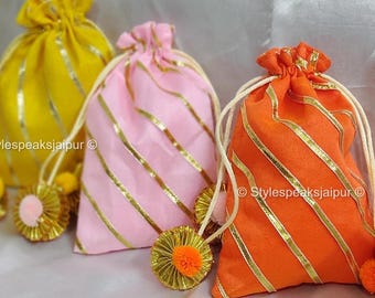 May include: Three drawstring bags in yellow, pink, and orange, each adorned with gold stripes and a matching tassel. The bags are tied with white cords and sit on a soft, white surface. The text "Stylespeaksjaipur" is visible.