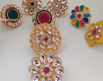 May include: A collection of gold-toned rings with floral designs. Some rings feature red, green, and clear gemstones. One ring has a yellow enamel base. The rings are arranged on a white surface.