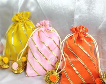 May include: Three colorful drawstring bags: yellow, pink, and orange. Each bag features gold diagonal stripes and a matching tassel. The bags are tied with white cords and sit on a white surface.