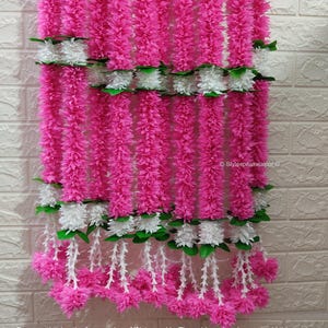 May include: A vibrant pink floral garland with white and green accents. The garland is composed of long, vertical strands of pink flowers, interspersed with sections of white flowers and green leaves. The text "Stylespeaksjaipur" is visible.