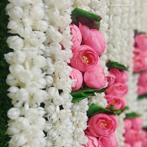 May include: Close-up of a floral garland featuring white and pink flowers. The garland is composed of white, bead-like flowers and clusters of pink roses with green leaves. The background is a blurred green.
