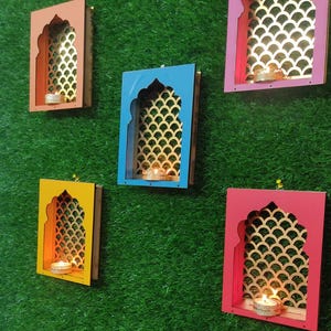 Puede incluir: Colección de coloridos candelabros montados en la pared. Cada soporte presenta un diseño de arco decorativo con un respaldo dorado estampado. Los soportes son en tonos rosa, azul, amarillo y naranja, con una pequeña vela dentro de cada uno.