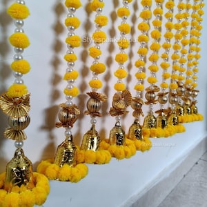 May include: A decorative garland featuring strands of yellow pom-poms, faux pearls, and gold-colored bells. The garland is suspended against a white wall. The text "Stylespeaksjaipur" is visible on the wall.
