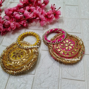 May include: Two decorative, round handbags. One is yellow with gold embellishments and a pearl-studded handle. The other is pink with gold accents and a pink and gold handle. Both have intricate designs and a textured appearance.
