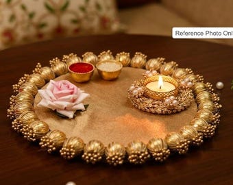 Può includere: Un vassoio decorativo circolare color oro con un bordo di perline metalliche dorate e accenti a forma di fiore. Il vassoio contiene una rosa artificiale rosa, due piccole ciotole dorate con polvere rossa e riso, e una candela accesa.