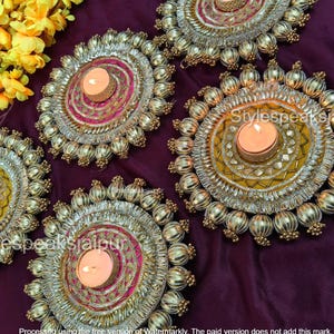 May include: Five decorative candle holders with gold and silver embellishments, each holding a lit candle. The holders have a circular design with beaded edges and a central area with pink and yellow accents. The background is a deep purple.