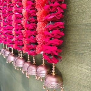 May include: A decorative garland featuring red roses and orange pom-poms, complemented by gold-toned bells. The floral arrangement is suspended, creating a decorative display. The bells have small dangling accents. The text "Stylespeaks Jaipur" is visible.