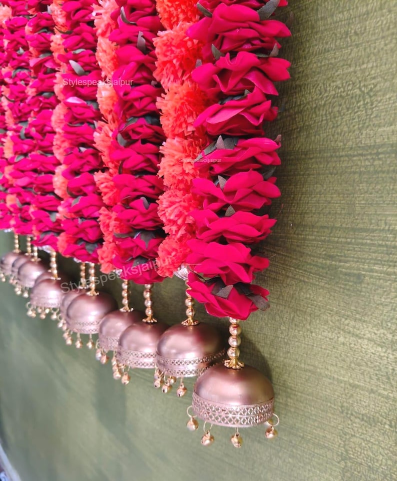 May include: A decorative garland featuring red roses and orange pom-poms, complemented by gold-toned bells. The floral arrangement is suspended, creating a decorative display. The bells have small dangling accents. The text "Stylespeaks Jaipur" is visible.