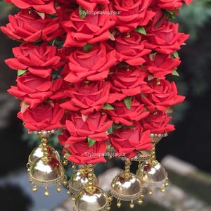 Puede incluir: Un adorno colgante decorativo que presenta múltiples capas de rosas artificiales de color rojo vibrante. El arreglo floral está acentuado con pequeñas cuentas doradas y termina en varios dijes de campana jhumka dorados pulidos. El texto Stylespeaksjaipur es visible.