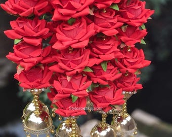 May include: A decorative hanging ornament featuring multiple layers of vibrant red artificial roses. The floral arrangement is accented with small gold-toned beads and terminates in several polished gold-toned jhumka bell charms. The text Stylespeaksjaipur is visible.