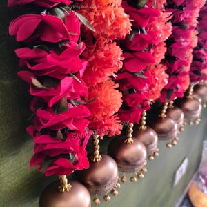 May include: A close-up of a decorative garland with bright red roses and orange pom-pom flowers. Bronze-coloured bell-shaped ornaments hang below, creating a colourful and festive display.