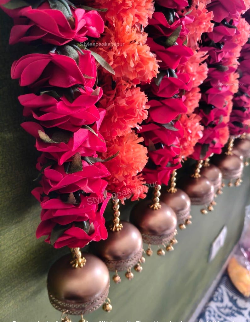 May include: A close-up of a decorative garland with bright red roses and orange pom-pom flowers. Bronze-coloured bell-shaped ornaments hang below, creating a colourful and festive display.