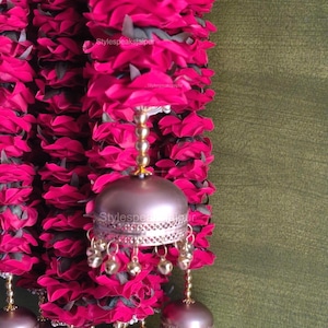 May include: A close-up of a garland with vibrant pink fabric flowers and decorative metal bells. The bells are a metallic bronze colour with small dangling gold-coloured bells. The garland is against a green background.