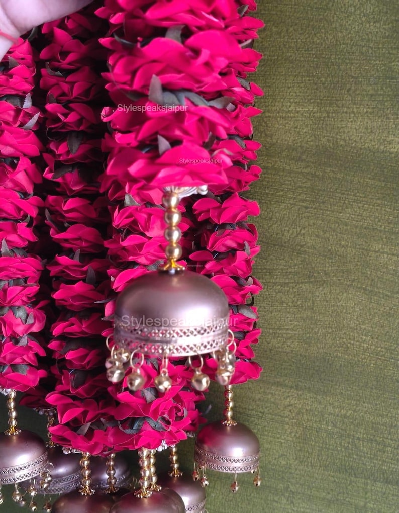 May include: A close-up of a garland with vibrant pink fabric flowers and decorative metal bells. The bells are a metallic bronze colour with small dangling gold-coloured bells. The garland is against a green background.