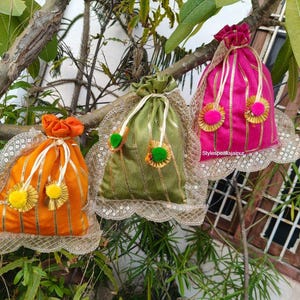 Può includere: Tre colorate borse con coulisse sono appese a un ramo di un albero. Le borse sono arancioni, verdi e rosa, con dettagli dorati e finiture decorative. Ogni borsa ha un design unico ed è impreziosita da nappe e dettagli floreali.