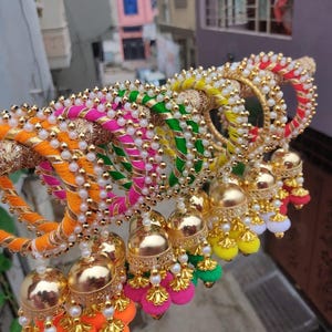 Puede incluir: Una colección de coloridas y ornamentadas pulseras y campanas decorativas. Las pulseras están envueltas en hilos vibrantes de color naranja, rosa, verde y amarillo, adornadas con cuentas blancas y detalles dorados. Cada campana tiene un acabado dorado y pompones coloridos.