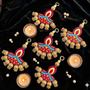 May include: Four decorative hanging ornaments shaped like traditional oil lamps, featuring magenta, cyan, and yellow patterns with gold-toned metallic beads. Three small glass tea light holders with lit candles are arranged among the ornaments.