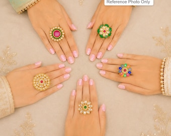 May include: Five gold-toned rings featuring various designs with colorful stones and pearls. One ring has a large pink center stone, another a green floral shape, one a multi-colored cluster, one a filigree pattern, and one a central green stone.