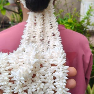 May include: A long, multi-strand garland of small, white, star-shaped jasmine flowers. The floral accessory is arranged in a dense, layered pattern, featuring a circular base that encircles a dark hair bun and long, cascading strands of white blossoms.