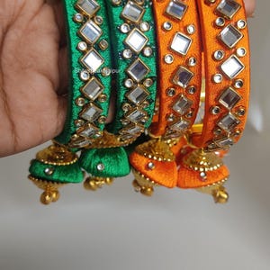 Puede incluir: Brazaletes de seda verdes y naranjas con detalles de espejo y colgantes en forma de campana. Los brazaletes están adornados con espejos cuadrados y adornos dorados. El texto "Stylespeaksjaipur" es visible en la imagen.