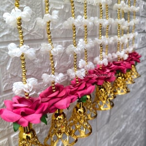 May include: Decorative garland featuring white floral accents, gold beads, and vibrant pink roses. Gold-colored bell-shaped ornaments hang below the roses. The text "Stylespeaksjaipur" is visible on the garland.