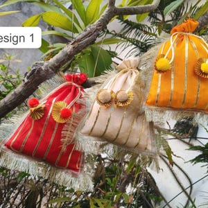 Puede incluir: Tres bolsas decorativas con cordón en rojo, beige y naranja, adornadas con rayas y ribetes dorados. Cada bolsa presenta una borla y detalles decorativos, colgando de una rama de árbol. El texto "Design 1" está en un recuadro blanco.