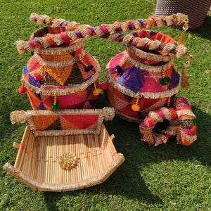 May include: A set of colourful, woven decorative items. Two round, lidded containers with fringed edges and tassel accents sit next to a rectangular tray and a circular ring. The items are made of woven materials in vibrant colours.
