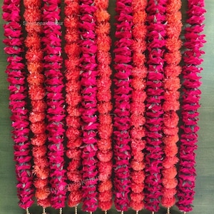 May include: A decorative floral garland in shades of red and orange, with gold-coloured bells. The garland is made up of alternating strands of deep red and orange flowers. The text "Stylespeaksjaipur" is visible on the garland.