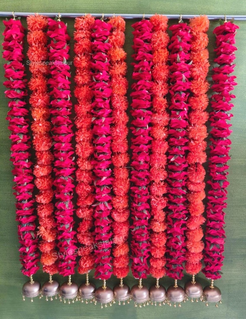 May include: A decorative floral garland in shades of red and orange, with gold-coloured bells. The garland is made up of alternating strands of deep red and orange flowers. The text "Stylespeaksjaipur" is visible on the garland.