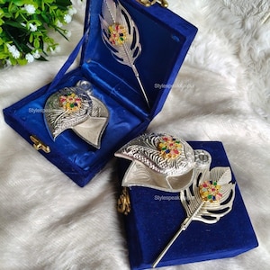 Puede incluir: Dos broches plateados en forma de pluma con detalles de piedras preciosas de colores, presentados en una caja de terciopelo azul. La caja está abierta, revelando el broche y un alfiler a juego en forma de pluma. El texto "Stylespeaksjaipur" es visible.