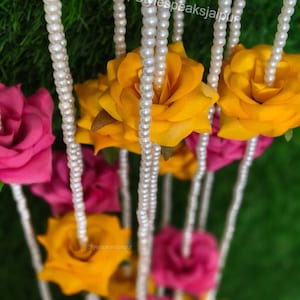 May include: A close-up of a floral garland featuring yellow and pink roses, interspersed with strands of white pearls. The text "Stylespeaksjaipur" is visible. The background is a blurred green.