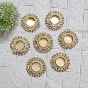 Pode incluir: Seis castiçais decorativos com um centro dourado e uma borda de contas brancas. A borda externa é adornada com pequenas contas douradas. Os castiçais são dispostos sobre uma superfície branca texturizada.