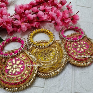 Könnte beinhalten: Drei verzierte, runde Handtaschen in Gelb und Pink mit goldenen Verzierungen und Perlenhenkeln. Die Taschen sind mit aufwendigen Mustern verziert und von rosa Blüten umgeben. Der Text "Stylespeaksjaipur" ist auf einer der Taschen zu sehen.