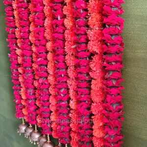 May include: Vertical rows of bright pink and orange flower garlands. The garlands are decorated with small, gold-coloured bells at the base. The background is a muted green, contrasting with the bright colours of the flowers.