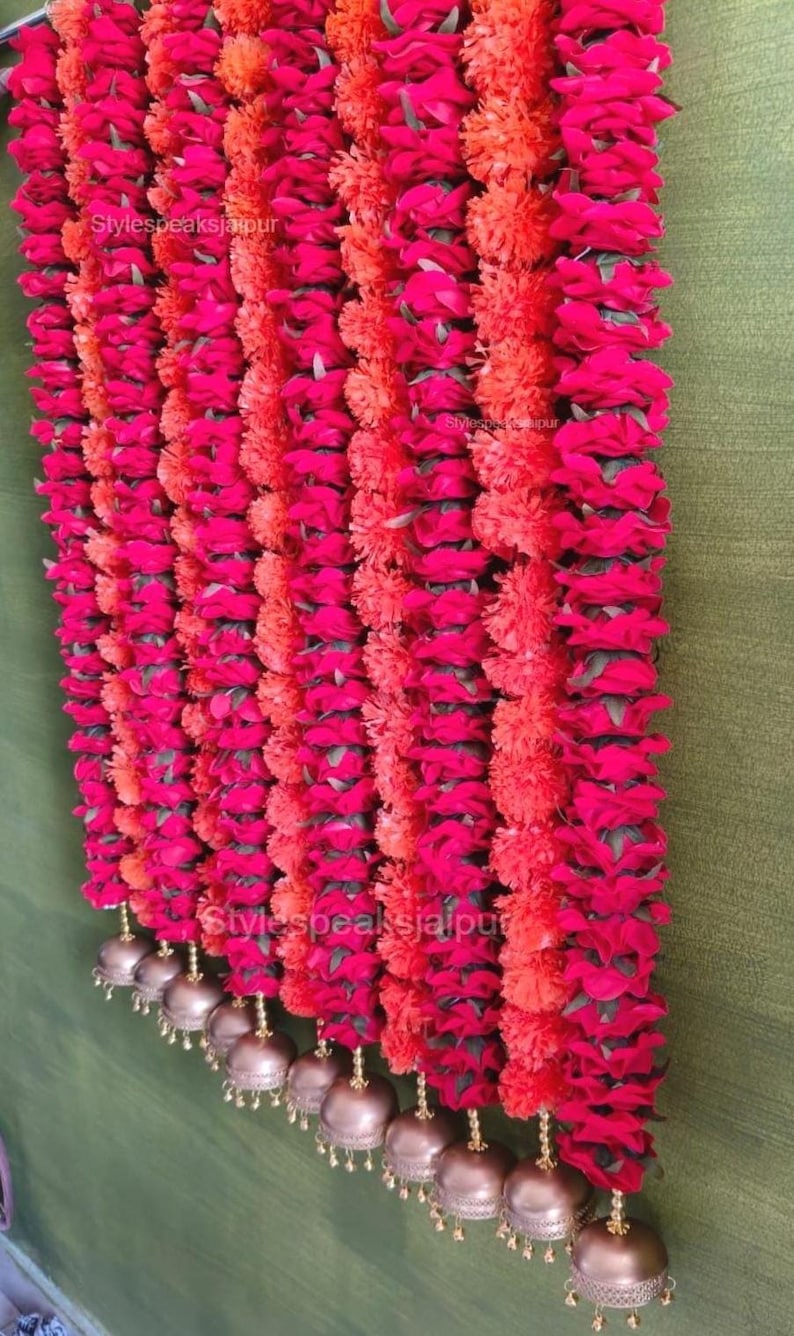May include: Vertical rows of bright pink and orange flower garlands. The garlands are decorated with small, gold-coloured bells at the base. The background is a muted green, contrasting with the bright colours of the flowers.