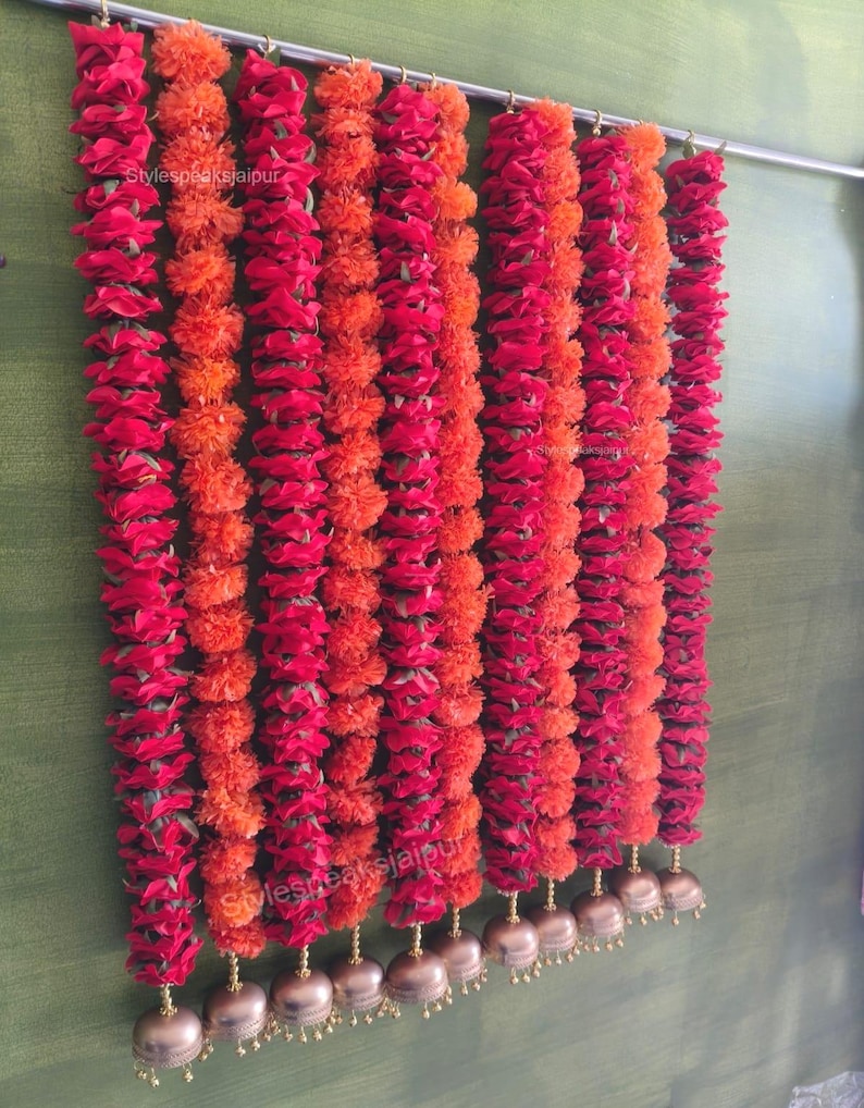 May include: A display of vibrant red and orange floral garlands hanging from a metal rod. The garlands are composed of densely packed flowers, alternating between red and orange hues. Gold-coloured bells dangle at the bottom of each garland. The text "Stylespeaksjaipur" is visible.