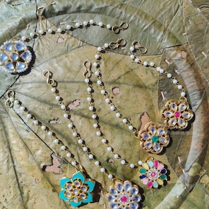 May include: An assortment of gold-toned, floral-shaped hair accessories with white pearl chains. The jewelry features colorful gemstones and a central design. The pieces are displayed on a textured, green-brown surface.
