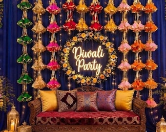 Könnte beinhalten: Ein Holzsofa mit rotem Samtsitz und fünf Zierkissen vor einem dunkelblauen Vorhang. Ein kreisförmiger Blumenkranz mit dem Leuchtschriftzug Diwali Party hängt über dem Sofa, flankiert von hängenden Quasten aus Stoff.
