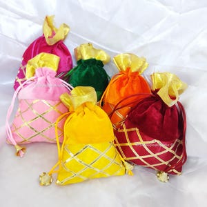 May include: A collection of colorful velvet drawstring bags in shades of pink, green, orange, yellow, and burgundy. Each bag features a gold metallic geometric pattern and a gold ribbon drawstring closure. The bags are arranged on a white surface.