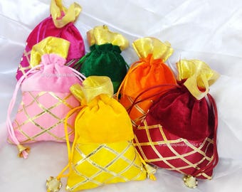 May include: A collection of colorful velvet drawstring bags in shades of pink, green, orange, yellow, and burgundy. Each bag features a gold metallic geometric pattern and a gold ribbon drawstring closure. The bags are arranged on a white surface.
