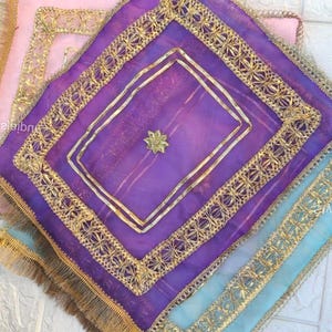 Może przedstawiać: Stos dekoracyjnych kwadratowych tekstyliów w odcieniach fioletu, różu i błękitu. Każdy materiał ma złote wykończenia i złotą ozdobę w kształcie gwiazdy. Krawędzie wykończone są złotymi frędzlami.