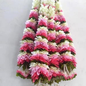 Puede incluir: Una guirnalda decorativa compuesta por flores en capas en tonos rosa, blanco y verde. La guirnalda presenta un diseño en cascada con una mezcla de texturas y una delicada hebra de perlas en la parte inferior. El texto "stylespeaksjaipur" es visible.