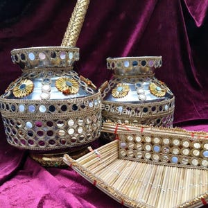 Puede incluir: Jarr&oacute;n decorativo plateado con intrincados adornos dorados y espejados. Los jarrones presentan detalles florales y van acompa&ntilde;ados de una bandeja de bamb&uacute;, todo sobre un fondo burdeos. Conjunto decorativo.