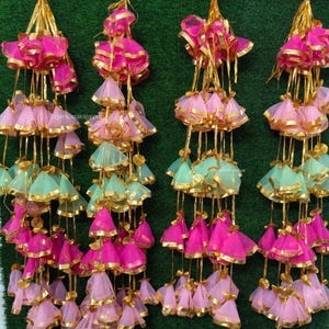 Peut inclure: Quatre guirlandes de pompons décoratifs roses et vert menthe, avec des bordures dorées. Chaque guirlande comporte plusieurs niveaux de petits ornements en forme de cloche, suspendus à des cordons dorés. Le fond est vert uni.