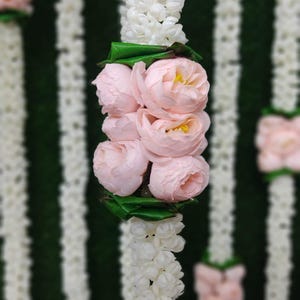 May include: Vertical image of a floral garland with white and pink flowers. The garland is composed of white, small, round flowers and clusters of pink roses with green leaves. The background is a dark green and white striped pattern.