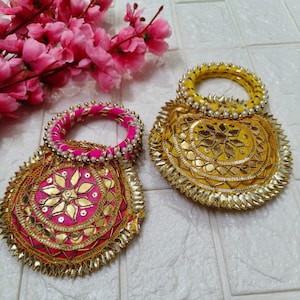 Puede incluir: Dos bolsos redondos y decorativos. Uno es rosa con adornos dorados y un asa con cuentas. El otro es amarillo con detalles dorados y un asa con cuentas. Ambos tienen un diseño floral y un borde decorativo. Flores rosas en el fondo.