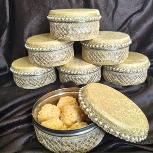 May include: A collection of decorative, round, lidded boxes with a gold and cream color scheme. The boxes are adorned with lace, sequins, and pearl trim. One box is open, revealing a serving of yellow, spherical food.