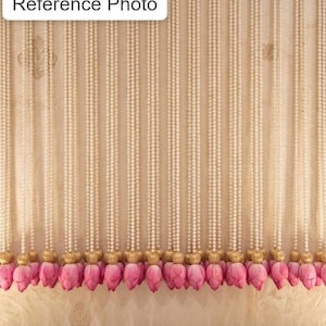 May include: A decorative curtain of white pearl strands, each ending with a gold-coloured accent and a cluster of pink lotus flower buds. The background is a light beige fabric with subtle patterns.